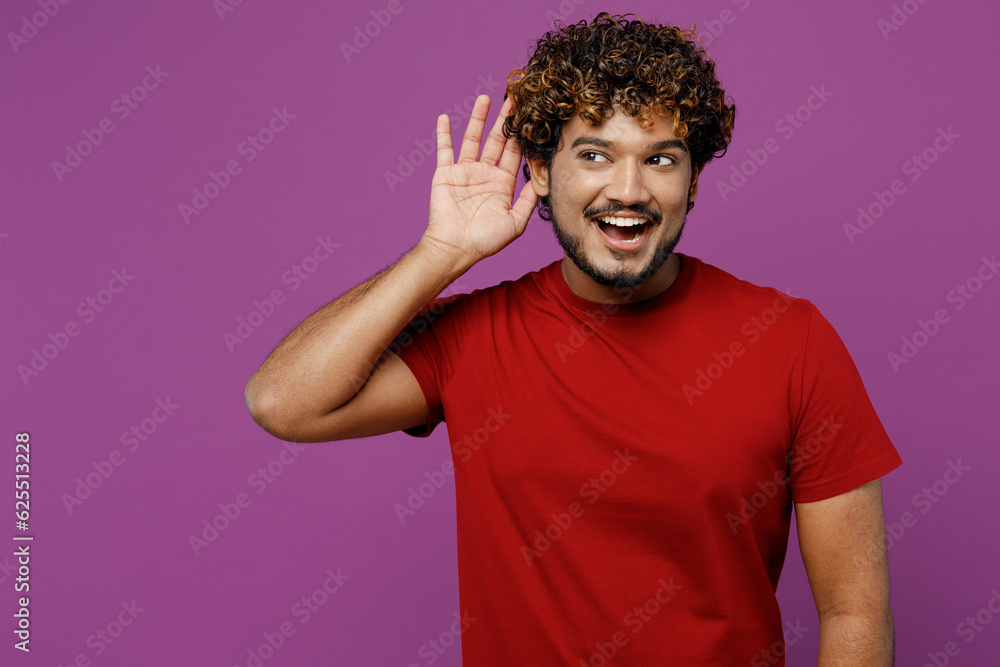 Young curious nosy smiling fun happy Indian man he wear red t-shirt casual clothes try to hear you overhear listening intently isolated on plain purple background studio portrait. Lifestyle concept. Young curious nosy smiling fun happy Indian man he wear red t-shirt casual clothes try to hear you overhear listening intently isolated on plain purple background studio portrait. Lifestyle concept.