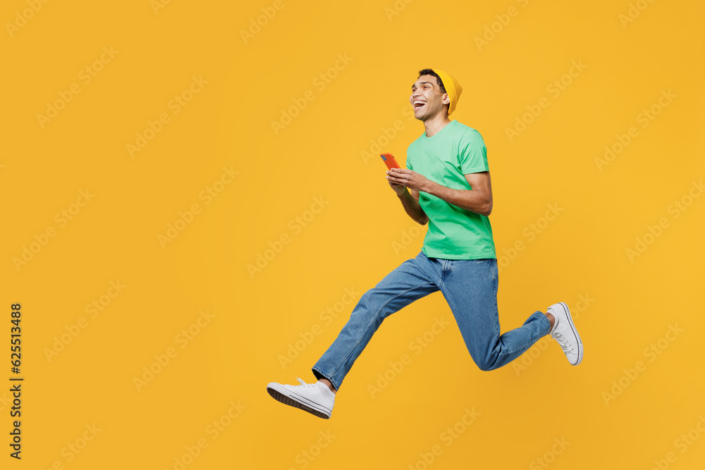 Full body side view young man of African American ethnicity he wears casual clothes green t-shirt hat jump high hold in hand use mobile cell phone run fast isolated on plain yellow background studio.