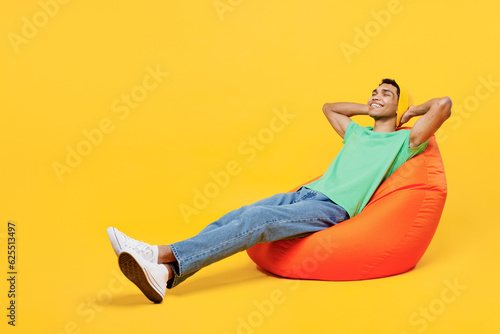 Full body young man of African American ethnicity he wears casual clothes green t-shirt hat sit in bag chair hold hands behind neck close eyes isolated on plain yellow background. Lifestyle concept.