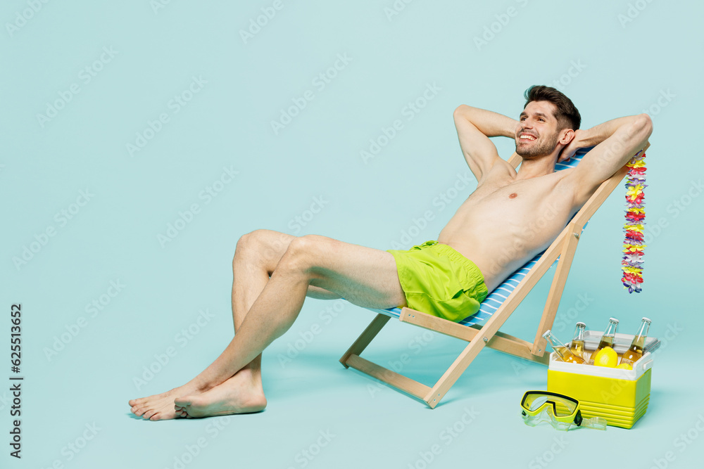 Obraz premium Full body side view young sexy man wear green shorts swimsuit relax near hotel pool sit in deckchair hold hands behind neck isolated on plain blue background. Summer vacation sea rest sun tan concept.