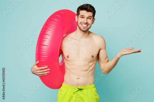 Young man wear green shorts swimsuit relax near hotel pool hold inflatable rubber ring shrugging shoulders spread hand isolated on plain blue cyan background. Summer vacation sea rest sun tan concept.