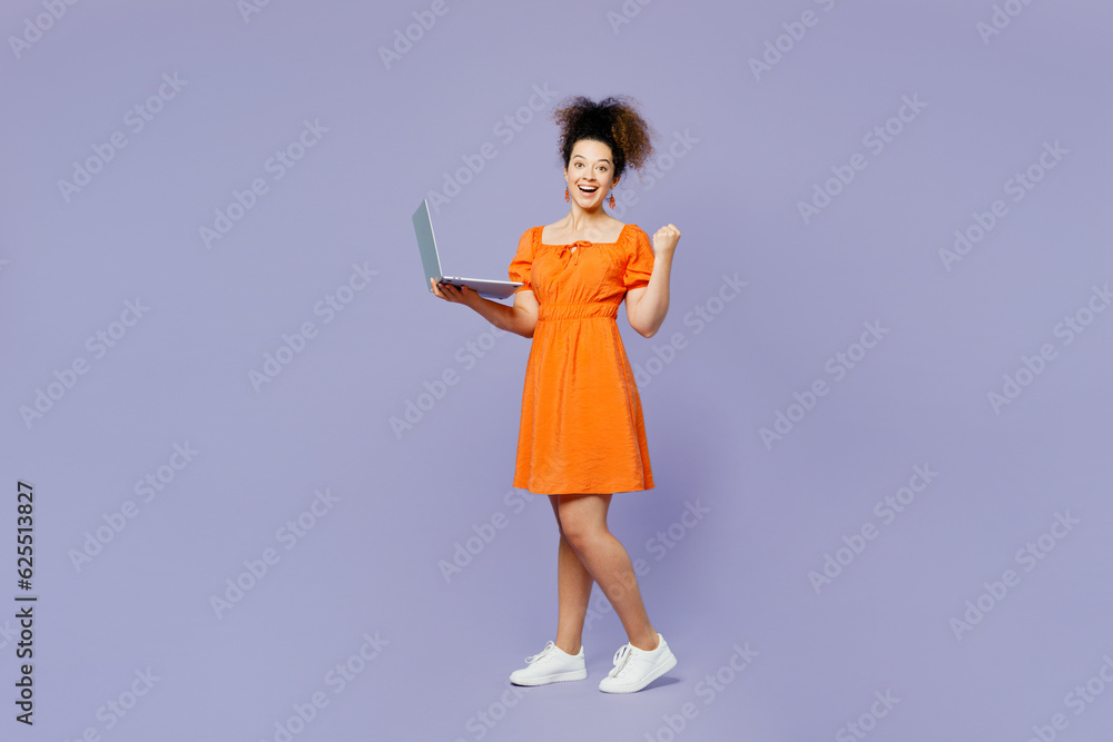 Full body side view young latin woman she wear orange blouse casual clothes hold use work on laptop pc computer do winner gesture isolated on plain pastel purple background studio. Lifestyle concept. Full body side view young latin woman she wear orange blouse casual clothes hold use work on laptop pc computer do winner gesture isolated on plain pastel purple background studio. Lifestyle concept.