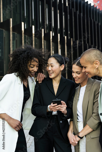 Diverse businesswomen group looking a phone and laughing standing in the street. Casual meeting between young females executives checking their smartphone outisde.