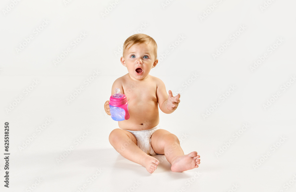 Cute, adorable baby, toddler in diaper sitting and eating, drinking water from bottle against white studio background. Concept of childhood, newborn lifestyle, happiness, care. Copy space for ad Cute, adorable baby, toddler in diaper sitting and eating, drinking water from bottle against white studio background. Concept of childhood, newborn lifestyle, happiness, care. Copy space for ad