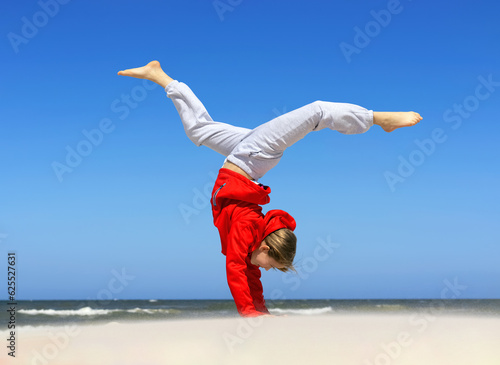 A young sporty girl in a red jacket does somersaults with raised legs against the background of the sea. Selective focus. Blurred background