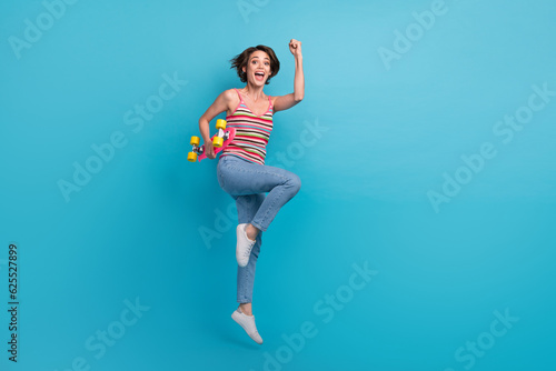 Fototapeta Naklejka Na Ścianę i Meble -  Full body photo of overjoyed nice lady jumping hold skateboard raise fists empty space isolated on blue color background
