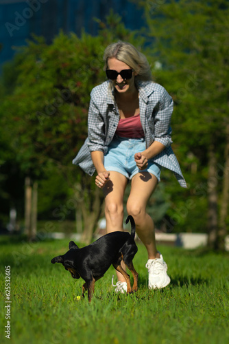 Young happy blonde woman is playing yellow ball with her black Toy Terrier dog in the park on the grass on a sunny day. Fun games with pets in the fresh air. Vertical photo
