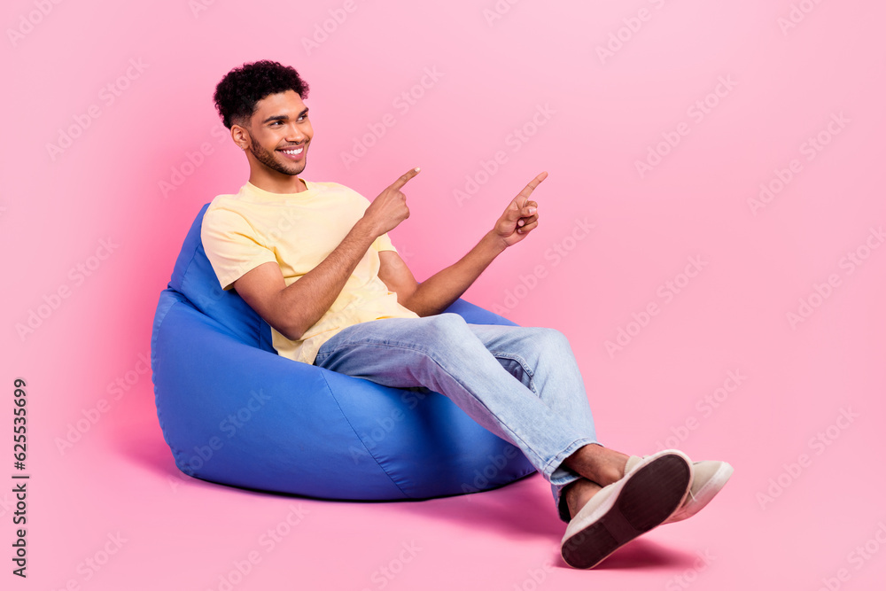 Full body photo of young chilling guy sitting pouf point fingers empty space recommend entertainment isolated on pink color background Full body photo of young chilling guy sitting pouf point fingers empty space recommend entertainment isolated on pink color background