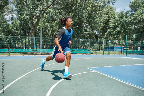 Dynamic image of young man, basketball player in blue uniform, training outdoors on playground on warm sunny day. Concept of professional sport, competition, hobby, game, active lifestyle © master1305