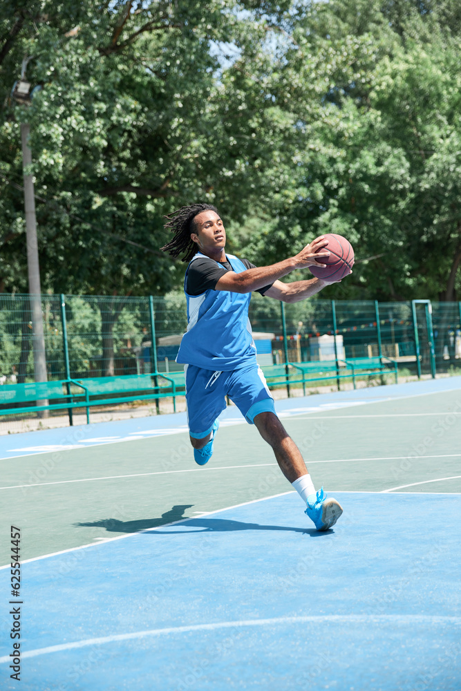 Full-length image of competitive young man in blue uniform playing basketball, throwing ball. Outdoor sportsground. Concept of professional sport, competition, hobby, game, active lifestyle