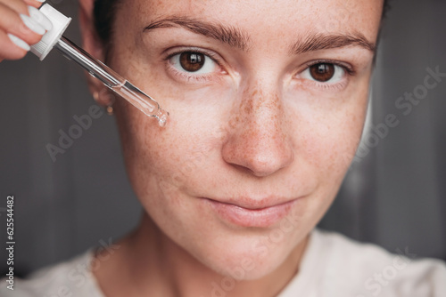 Young woman with no make up applying transperent serum on her face, brown eyes looking at the camera 