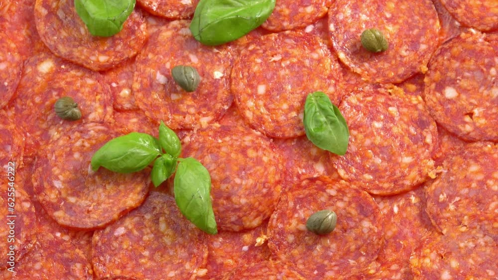 Juicy sliced pepperoni sausage with fresh basil and capers rotating as background for restaurant, food video, food porn. Smoked sausage slices for pizza Juicy sliced pepperoni sausage with fresh basil and capers rotating as background for restaurant, food video, food porn. Smoked sausage slices for pizza