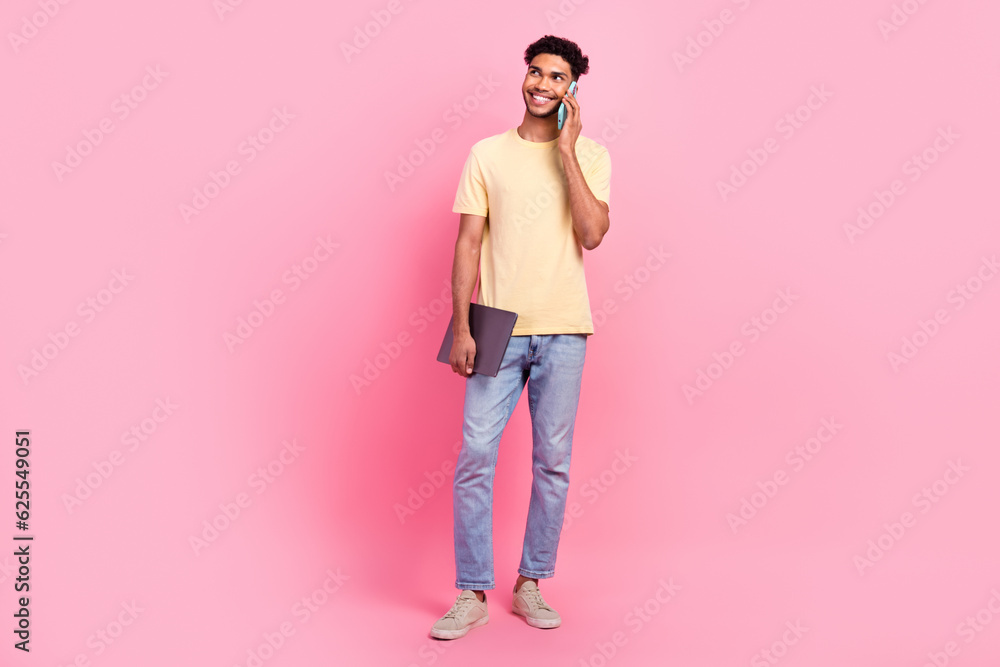 Full length photo of cool guy wear yellow shirt denim pants hold laptop look empty space talk on phone isolated on pink color background Full length photo of cool guy wear yellow shirt denim pants hold laptop look empty space talk on phone isolated on pink color background