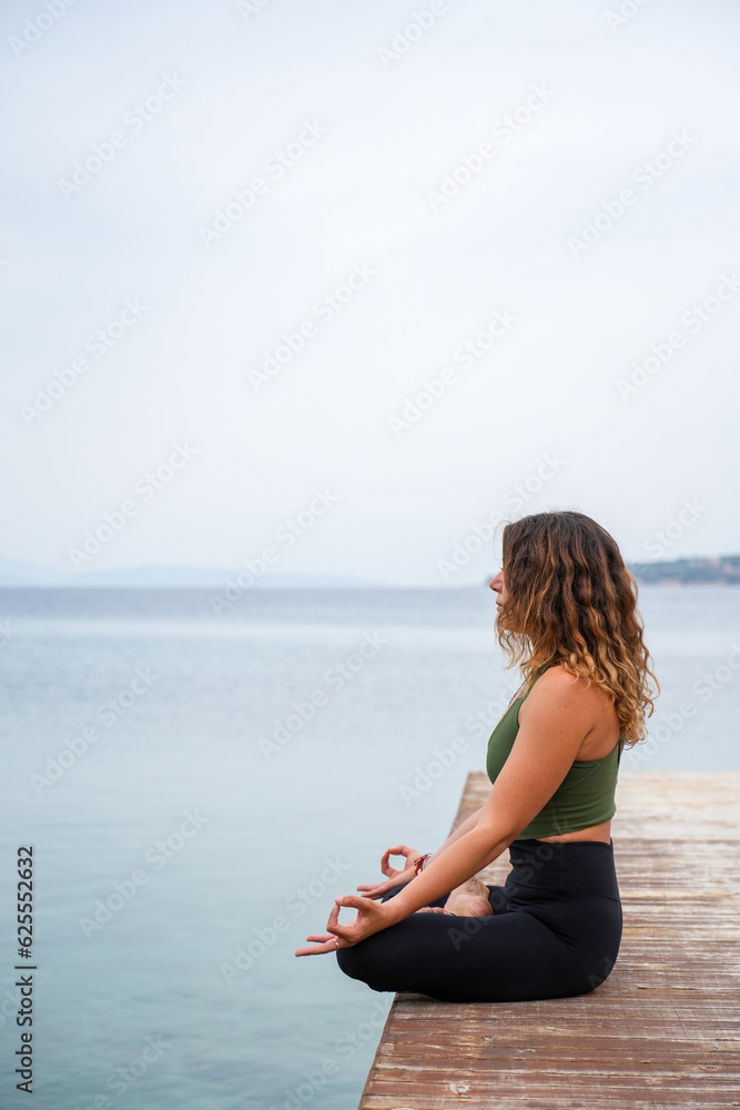 Woman practicing yoga by the sea on a deck - Padmasana / Lotus pose / meditation sitting - chin mudra
