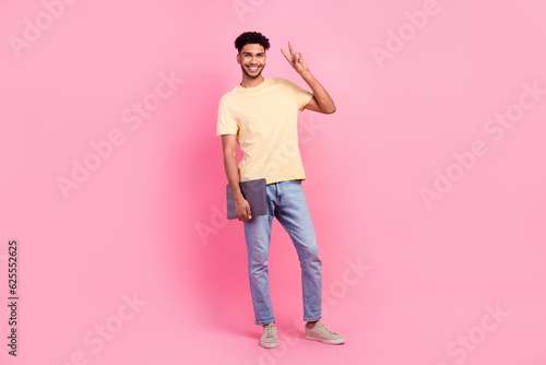 Full size photo of optimistic funny nice man wear yellow t-shirt jeans showing v-sign holding laptop isolated on pink color background