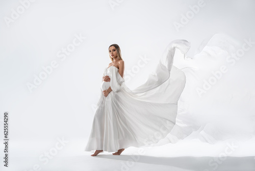 Delicate Pregnant Woman holding her Belly wearing white, long Dress. Happy mom to be. A lot of copy space.