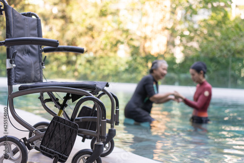 Selective focus off wheelchair background of physical therapist helping senior woman in aqua, old women and mature doing aqua aerobics exercise in swimming pool, Elderly sports, and active lifestyle.