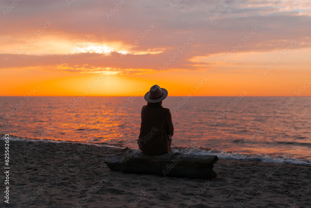 Back view of lonely adult thoughtful woman in coat and hat sitting on og on seashore and looking at sea sunset, mood. Unrecognizable pensive senior lady alone in nature in evening Back view of lonely adult thoughtful woman in coat and hat sitting on og on seashore and looking at sea sunset, mood. Unrecognizable pensive senior lady alone in nature in evening