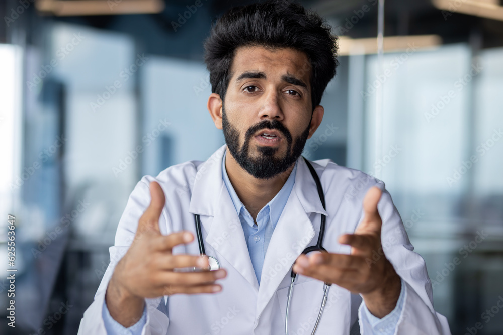 Online medical consultation. Portrait of a serious young Indian male doctor sitting in the office at the table talking, consulting the camera, gesturing with his hands Online medical consultation. Portrait of a serious young Indian male doctor sitting in the office at the table talking, consulting the camera, gesturing with his hands