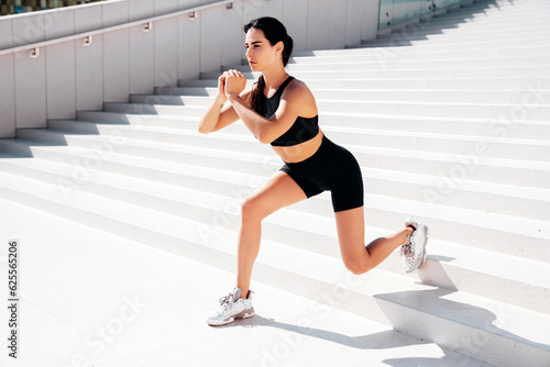 Fitness woman in sports clothing. Sexy young beautiful model athlete doing fitness workout. Female making exercises in the street at sunny day. Stretching out before training. Makes lunges at stairs