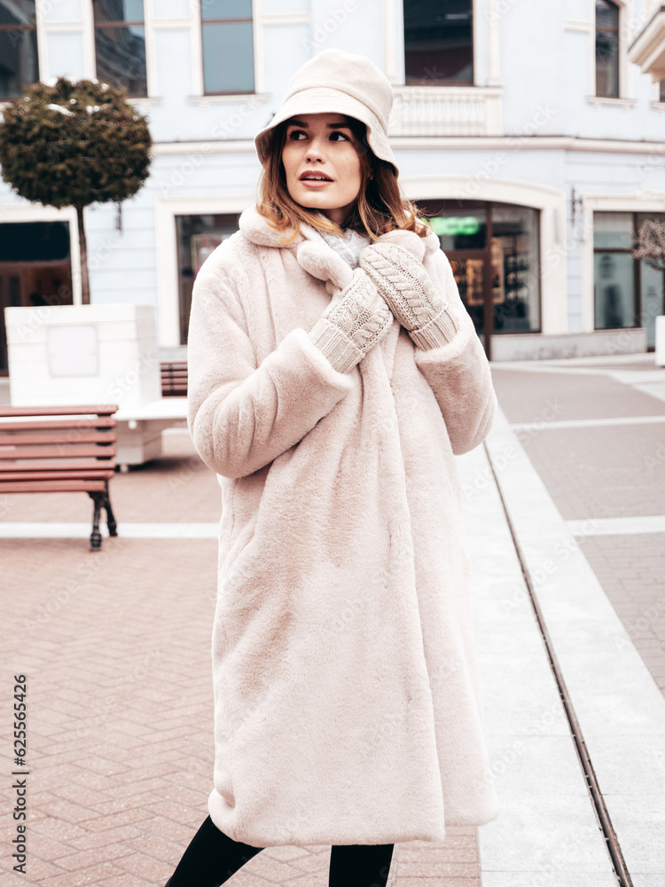 Outdoor fashion portrait of young beautiful smiling lady wearing trendy white faux fur coat. Stylish woman posing in the street in winter. Cheerful and happy model in panama. Cold autumn weather