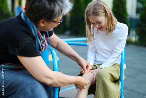 A mature woman patches up a wound on a child's leg at the playground.