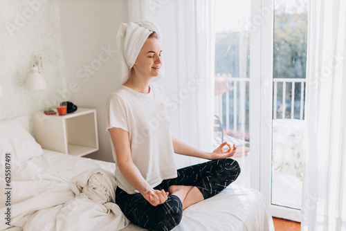 Concentrated young woman doing yoga, sitting in lotus pose on the bed. Wellbeing, healthy lifestyle concept