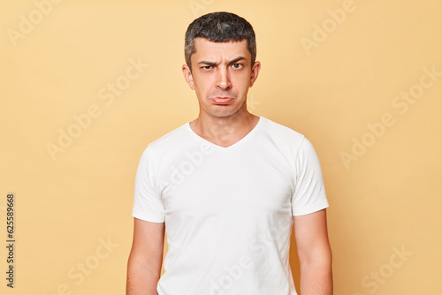 Unhappy offended man wearing white casual t-shirt standing isolated over beige background looking at camera with pout lips expressing sorrow and sadness.