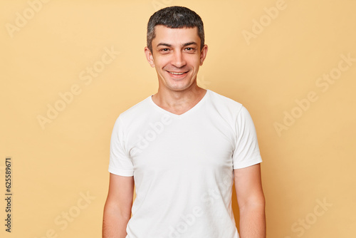 Smiling handsome man wearing white casual t-shirt standing isolated over beige background looking at camera being in good mood expressing positive emotions.