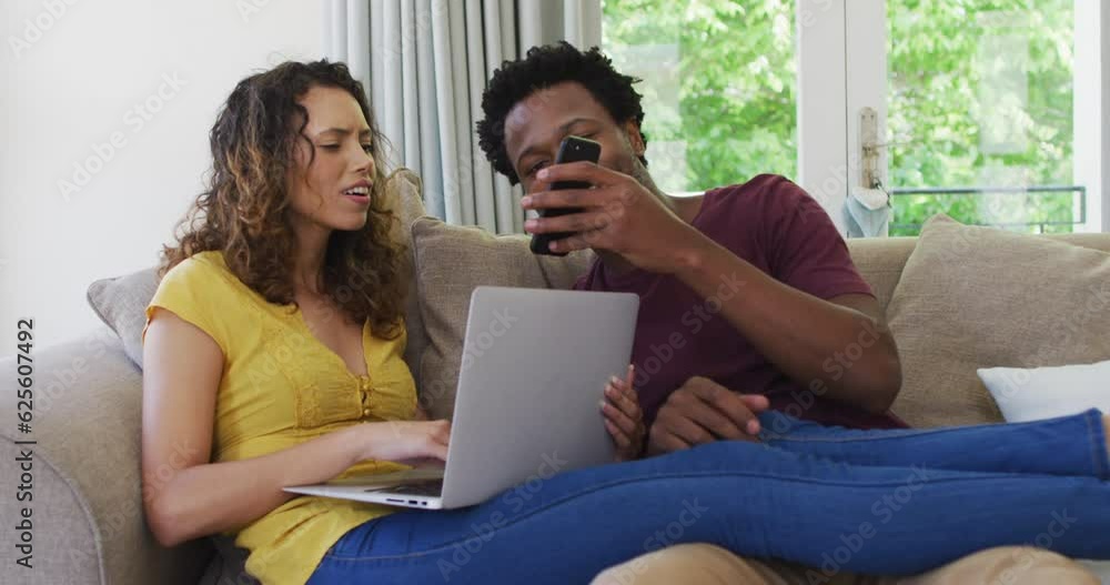 Happy biracial couple sitting on sofa with laptop and smartphone and laughing Happy biracial couple sitting on sofa with laptop and smartphone and laughing