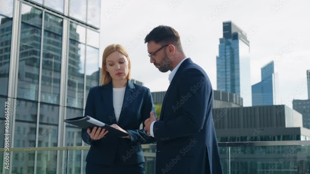Gimbal shot two business partners caucasian man woman in formal suits standing outdoors office building terrace in downtown city discussing upcoming project and deals looking at paper documents folder Gimbal shot two business partners caucasian man woman in formal suits standing outdoors office building terrace in downtown city discussing upcoming project and deals looking at paper documents folder