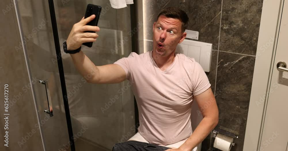 Male person takes funny selfie sitting on toilet bowl in light bathroom. Man poses for photo on mobile phone in premise of apartment slow motion Male person takes funny selfie sitting on toilet bowl in light bathroom. Man poses for photo on mobile phone in premise of apartment slow motion