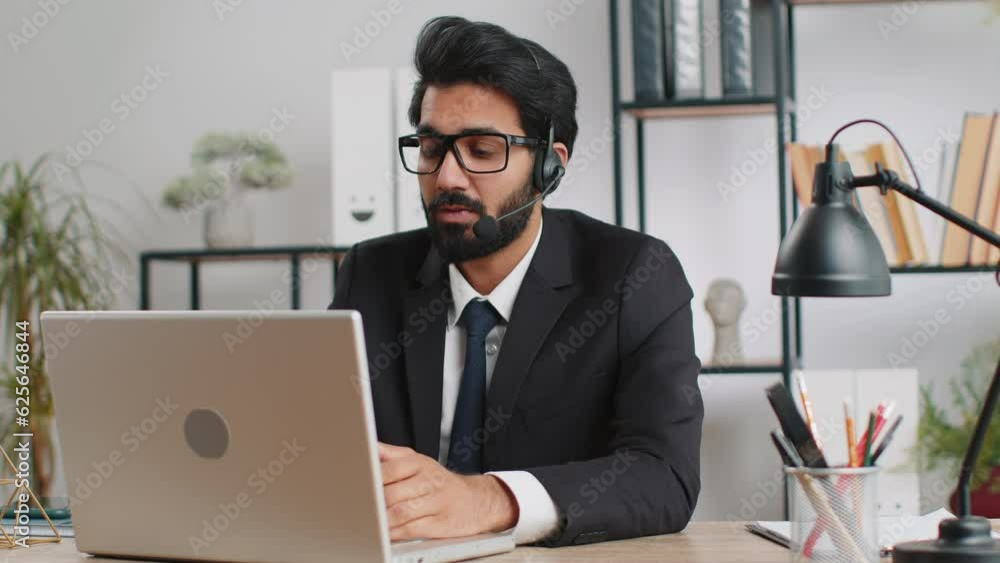 Indian business man working on laptop wearing headset, freelance worker call center or support service operator helpline, having pleasant talk with client or colleague communication support at office Indian business man working on laptop wearing headset, freelance worker call center or support service operator helpline, having pleasant talk with client or colleague communication support at office