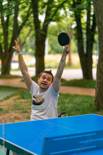 An inclusive disabled man with a racquet in his hand celebrates winning a game of ping pong against the backdrop of a city park © Guys Who Shoot