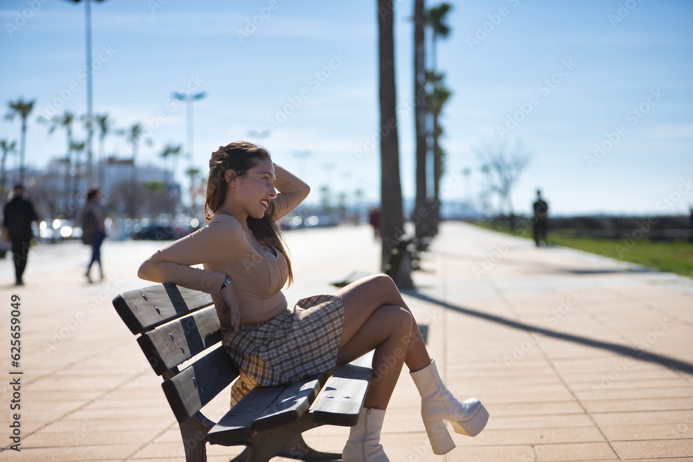Attractive young woman with blue eyes, sweater, plaid skirt and white platforms, sitting on a bench, relaxed and looking at infinity. Concept of relaxation, tranquility, peace.
