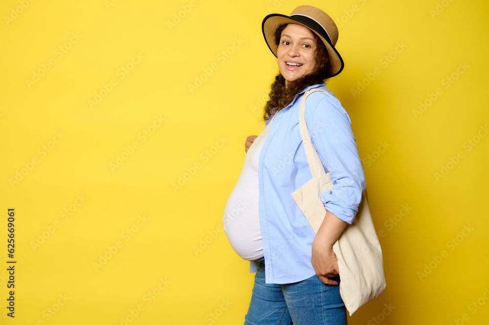 Portrait of a pregnant woman carrying an eco linen bag, smiling looking at camera, isolated on yellow studio background Portrait of a pregnant woman carrying an eco linen bag, smiling looking at camera, isolated on yellow studio background