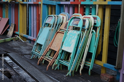Rusty painted folded metal chairs with paint peeling off leaning on colourful fence in a harbour with wooden floor in a outdoor environment. Wet wooden floor in an outdoor environment.