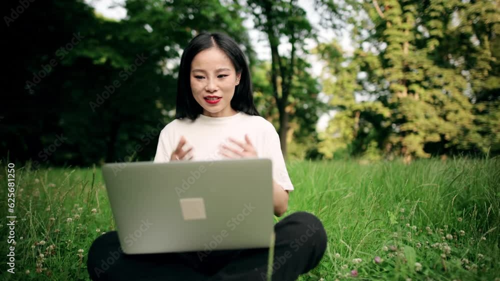 Pretty young asian woman sitting at park and having a video conference call on her laptop. Portrait of attractive girl sitting on grass using laptop outdoor