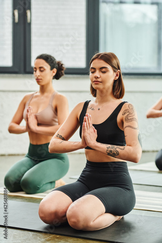 young tattooed woman sitting in thunderbolt pose and meditating with closed eyes and praying hands near multiracial girlfriend on blurred background, inner peace and body awareness concept