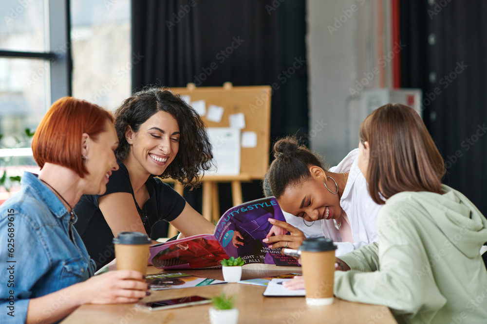 excited multicultural female friends reading beauty and style magazine and laughing near paper cups with takeaway drinks on table in women club, common interests and knowledge-sharing concept excited multicultural female friends reading beauty and style magazine and laughing near paper cups with takeaway drinks on table in women club, common interests and knowledge-sharing concept