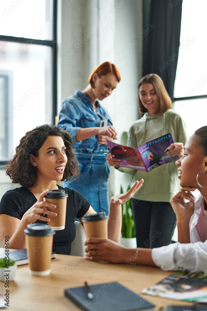 Obraz premium multiracial woman with takeaway drink gesturing and talking to african american girlfriend near members of interest club reading magazine on blurred background, solidarity and understanding concept