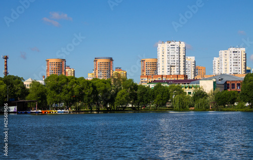 Beautiful urban landscape - modern multi-storey houses on the shore of a pond with reflection and green trees with lush foliage on a sunny summer day in Reutov and a space for copy