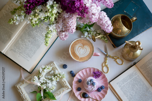 A cup of freshly brewed coffee with heart-shaped foam and spring lilac flowers, open books. Spring still life.