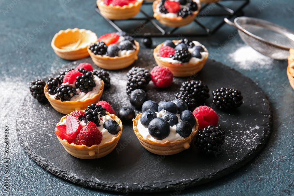 Board of tasty tartlets with whipped cream and berries on dark background
