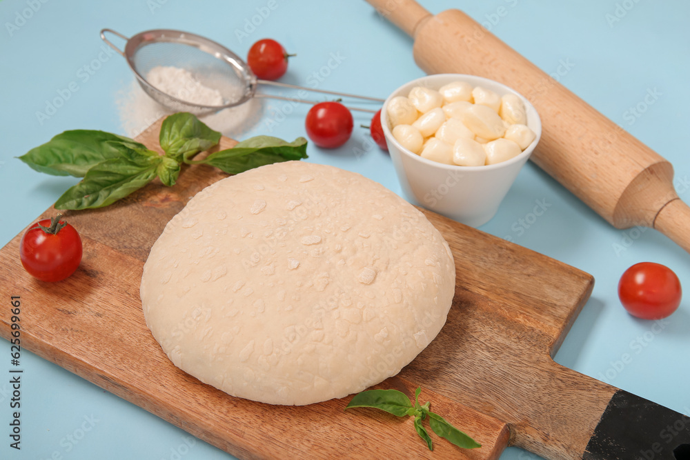 Wooden board with raw dough and ingredients for preparing pizza on blue background Wooden board with raw dough and ingredients for preparing pizza on blue background