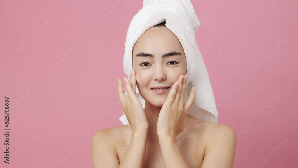 Asian young woman with a towel on her head looking at the camera, apply the cream, smiling on pink background. Beautiful female model with perfect clean fresh skin. Skin care treatment concept Asian young woman with a towel on her head looking at the camera, apply the cream, smiling on pink background. Beautiful female model with perfect clean fresh skin. Skin care treatment concept