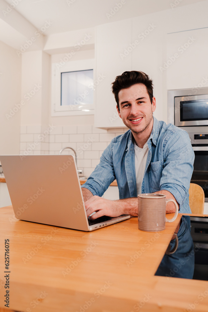Vertical portrait of handsome young man working remotely from home with a laptop looking at camera. Front view of happy entrepreneur male smiling using Internet with a computer sitting at home kitchen Vertical portrait of handsome young man working remotely from home with a laptop looking at camera. Front view of happy entrepreneur male smiling using Internet with a computer sitting at home kitchen