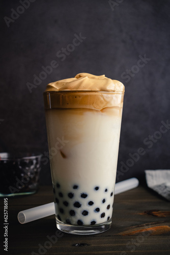 Iced Dalgona Coffee with Boba in a Pint Glass on a Dark Background: Cold dalgona coffee with milk and tapioca pearls shown with a boba straw