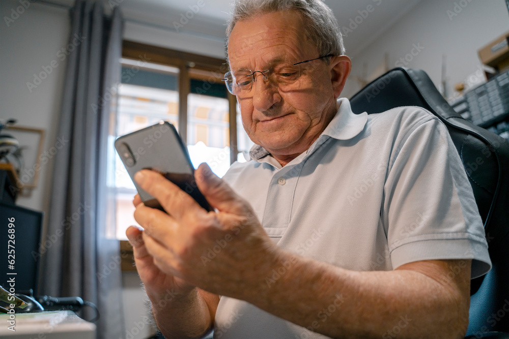 Smiling senior man holding mobile phone, chatting, text message, sitting in working workshop at home Smiling senior man holding mobile phone, chatting, text message, sitting in working workshop at home