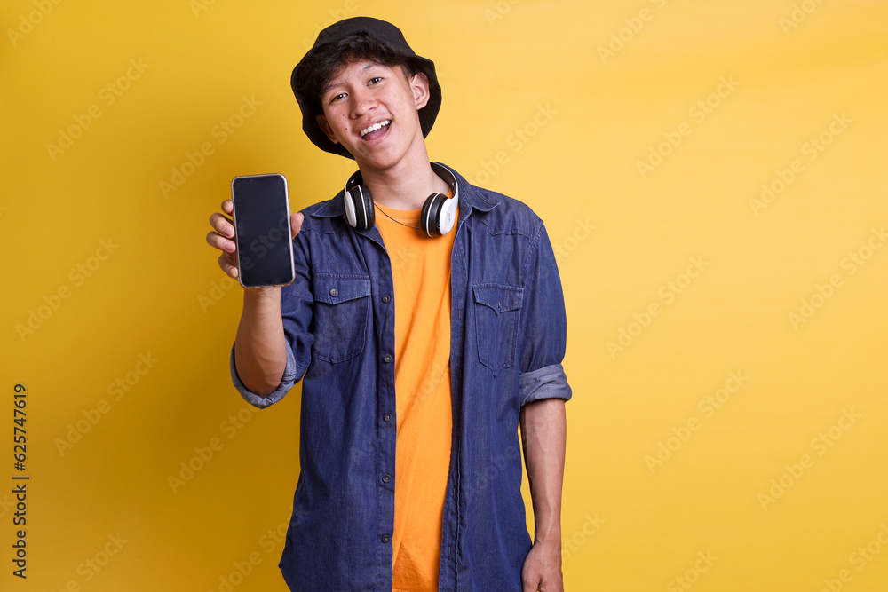 Young cheerful male student in casual clothes, bucket hat with headphones on his neck showing blank screen smartphone mockup over yellow backgrounds Young cheerful male student in casual clothes, bucket hat with headphones on his neck showing blank screen smartphone mockup over yellow backgrounds